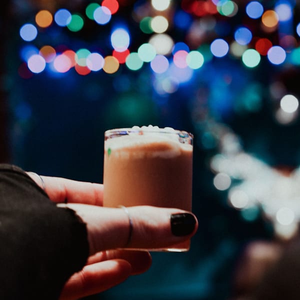 Hand holding a small glass of creamy beverage with white foam, against blurred colorful lights.