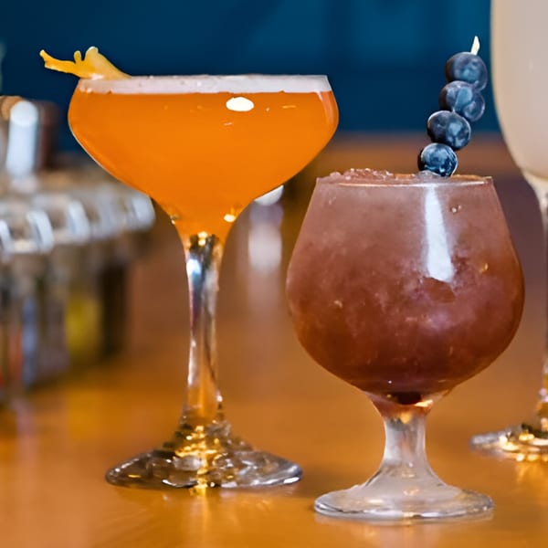 Two cocktails on a wooden bar, one orange with a citrus peel, the other purple with blueberries.