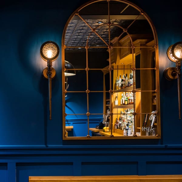 An arched mirror on a dark blue wall reflects a well-stocked bar with bottles and taps.