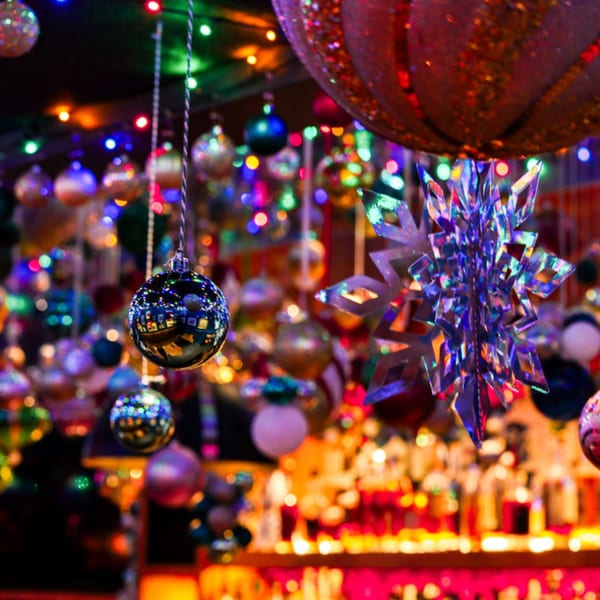 Colorful Christmas ornaments and string lights hang above a blurred bar with bottles and glowing lights.
