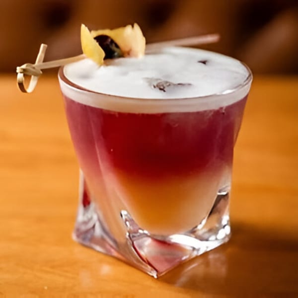 A layered cocktail with red and orange liquid, topped with white foam and a lemon peel garnish.