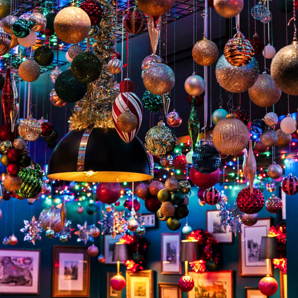 Numerous colorful Christmas ornaments and string lights hang from the ceiling, illuminating a dark room.