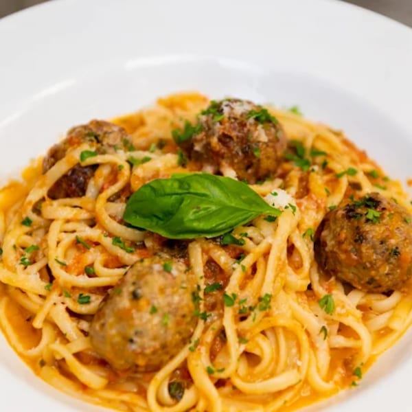 Linguine pasta with meatballs, tomato sauce, parmesan cheese, and a basil leaf on a white plate.