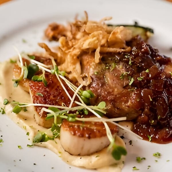 Seared scallops and braised short rib with crispy onions, microgreens, and a creamy sauce on a white plate.